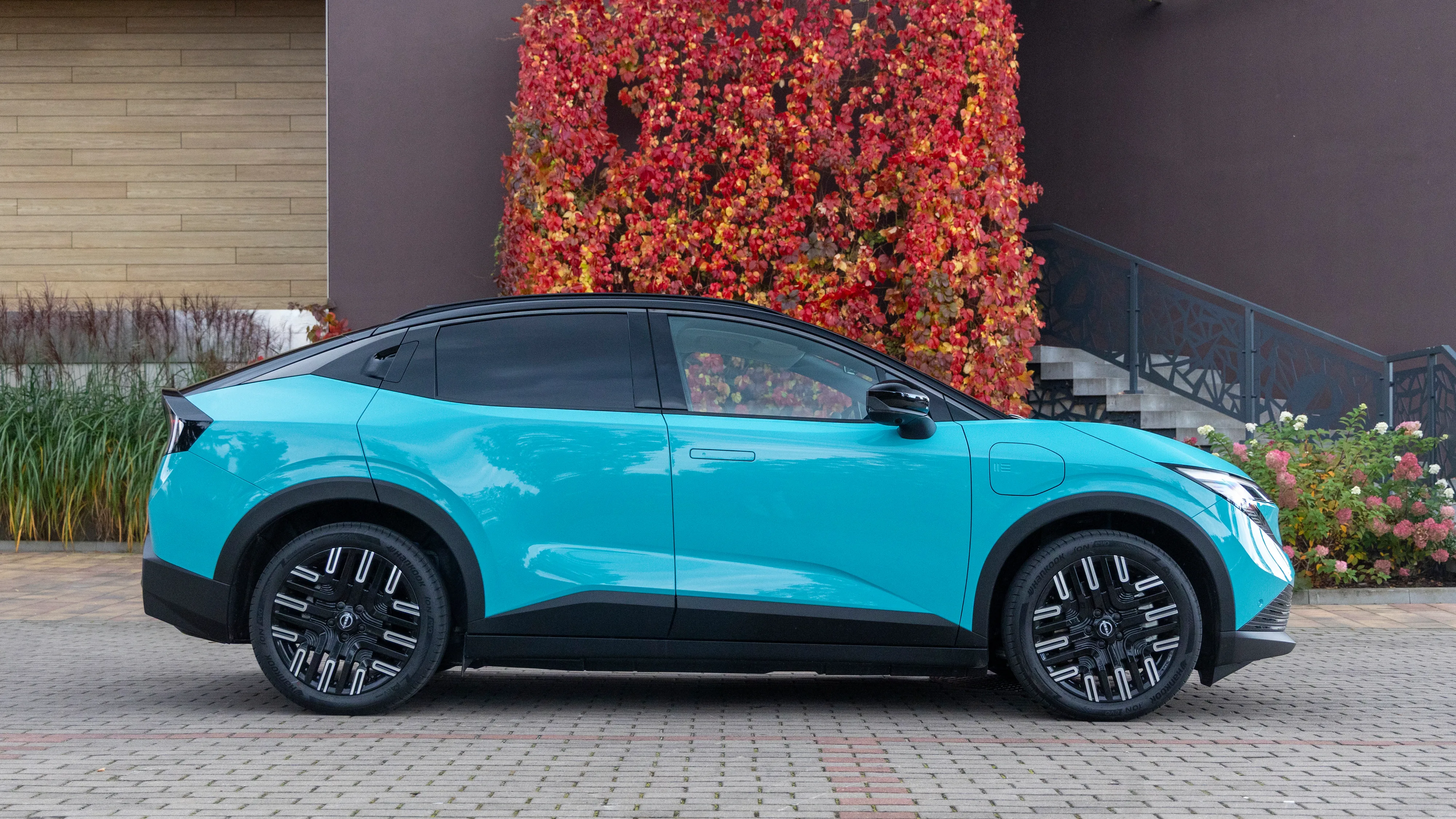 Turkusowy Nissan LEAF 2026 z czarnym dachem widziany z boku, zaparkowany przed budynkiem z czerwonymi liśćmi