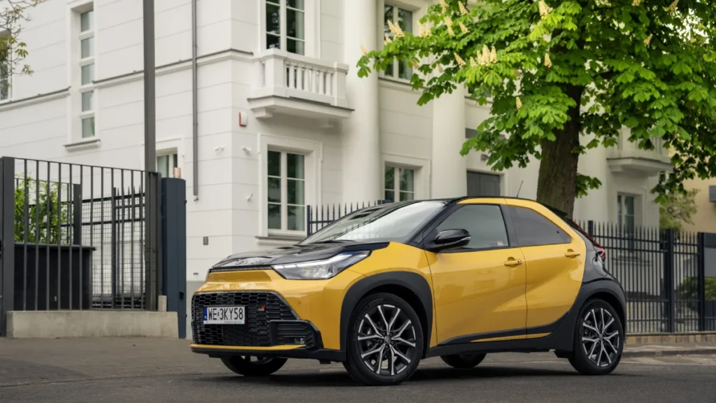 Żółta Toyota Aygo X Hybrid z czarnym dachem zaparkowana przed jasnym budynkiem