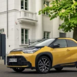 Żółta Toyota Aygo X Hybrid z czarnym dachem zaparkowana przed jasnym budynkiem